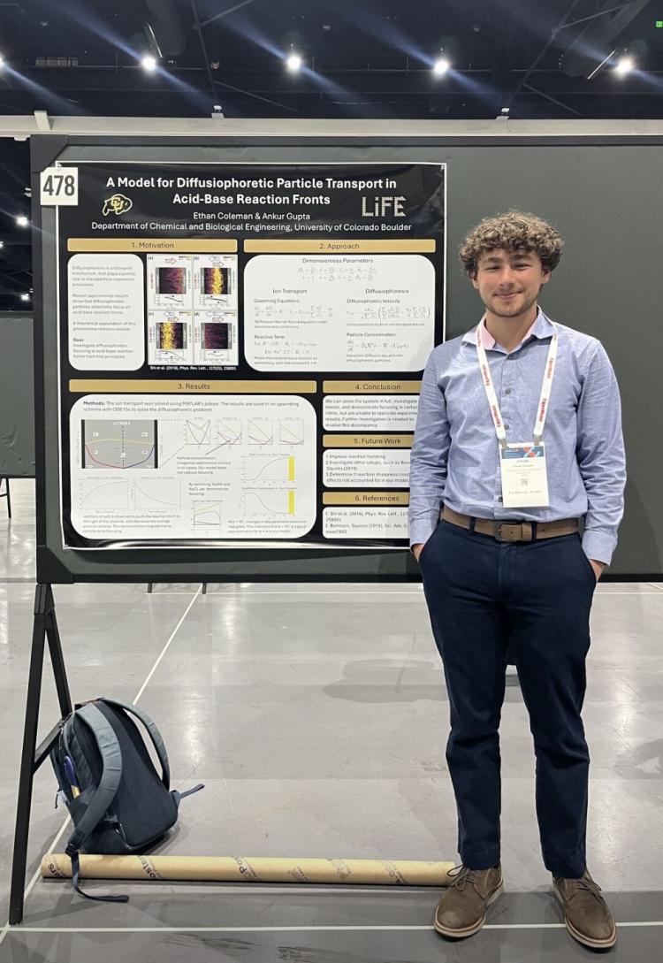 Ethan Coleman stands in front of a scientific research poster titled &ldquo;A Model for Diffusiophoretic Particle Transport in Acid-Base Reaction Fronts.&rdquo; He is smiling, wearing a light blue shirt and dark pants, with a conference badge around his neck. A backpack and poster tube rest on the floor beside the display in a large convention hall.