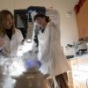 Two researchers in lab coats and safety goggles work with a cryogenic storage container emitting vapor. One holds a tray of samples above the container while the other holds the lid. The lab is equipped with microscopes, storage cabinets, and scientific equipment.