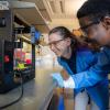Konane Bay peers inside of an instrument in her lab while a grad student looks on.