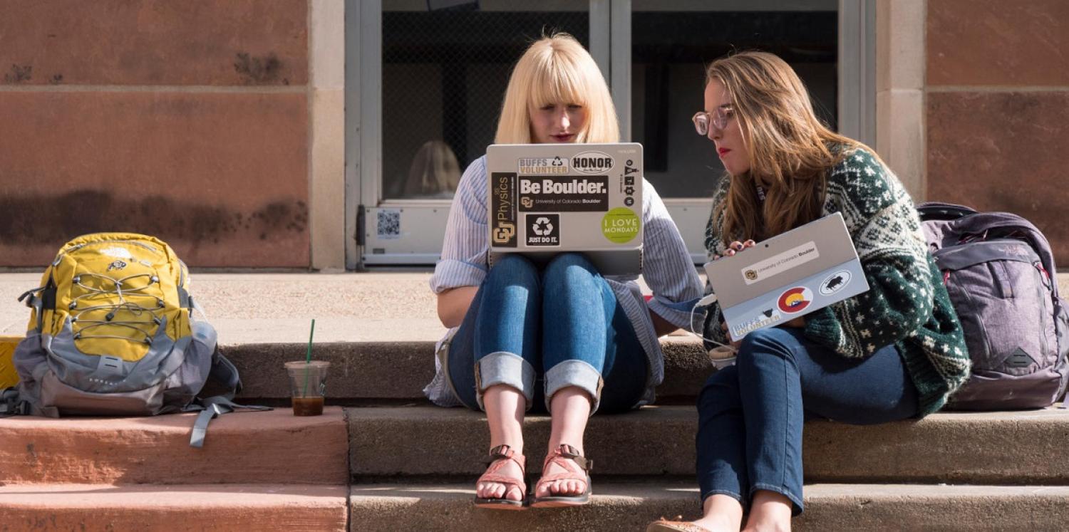 Two students sitting on the steps and looking at laptops