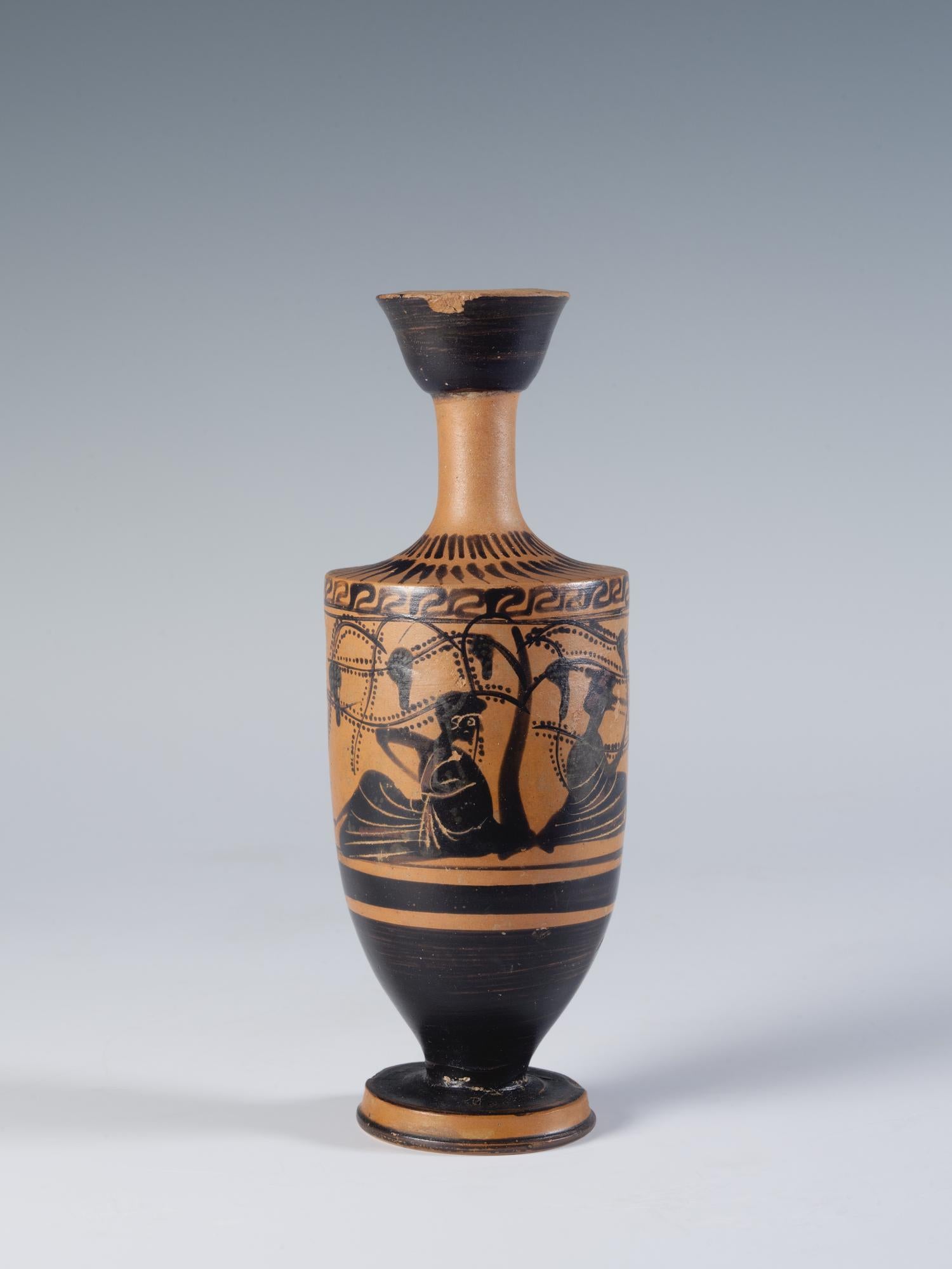 Photograph of a black-figure lekythos showing two human figures reclining beneath a productive grape arbor, from the side against a neutral gray background.