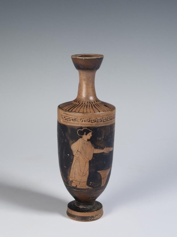 Photograph of red-figure lekythos against a neutral gray background, from the side and showing figure of a woman holding a dish over a wool basket.