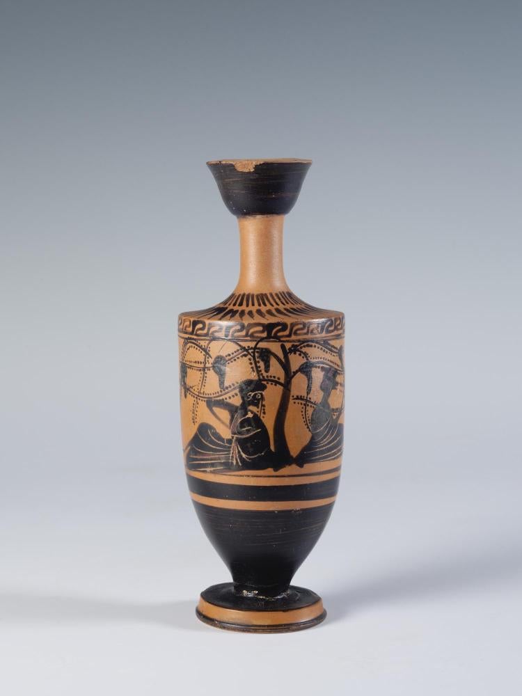 Photograph of a black-figure lekythos showing two figures reclining beneath a productive grape arbor, from the side against a neutral gray background.