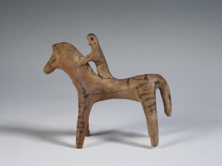 Photograph of a horse and rider figurine, facing toward viewer's left, from the side against a neutral gray background.