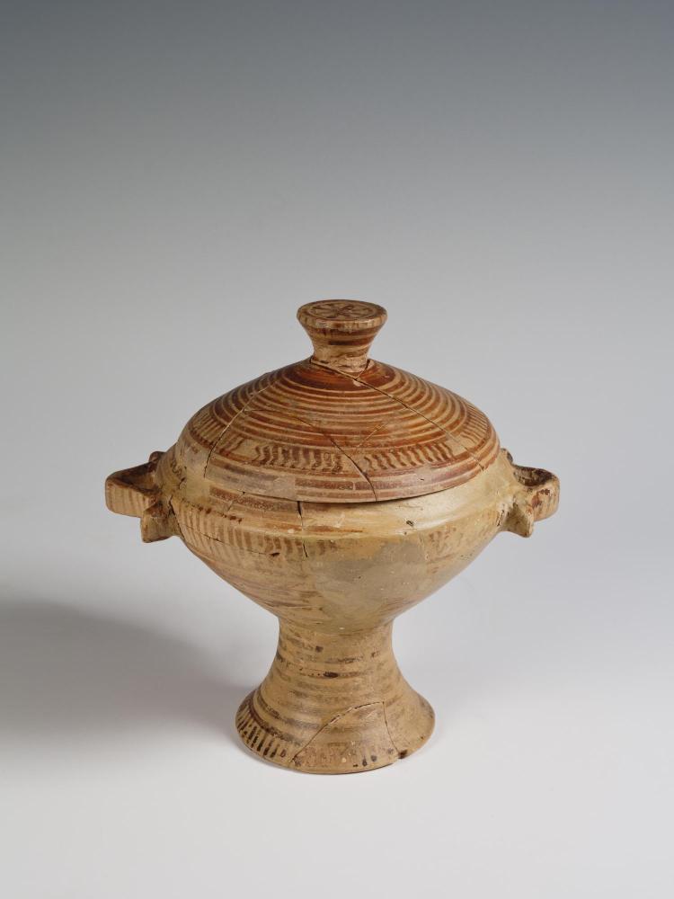 Photograph of a pedestalled skyphos with its lid intact, buff clay with red decoration, from the side against a neutral gray background.