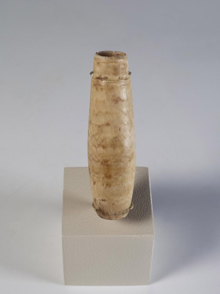 Photograph of a white stone alabastron with a tall, narrow body, supported on a regular whitish block and against a neutral gray background.