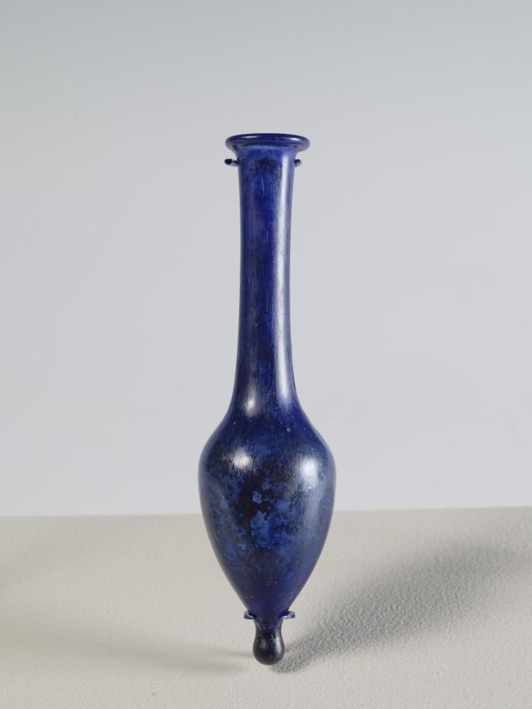 Photograph of a blue glass vessel with a pointed bottom, ovoid body, and tall, narrow neck that tapers slightly outward to a rounded mouth, from the side against a neutral background.