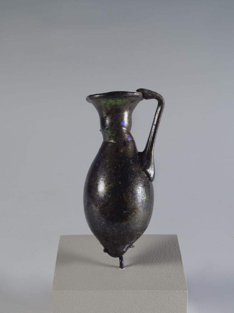 Photograph of a dark glass vessel with an oviod body that tapers to a point, a relatively short neck that tapers outward to the mouth, and a handle that connects the body to the mouth, from the side against a neutral gray background.
