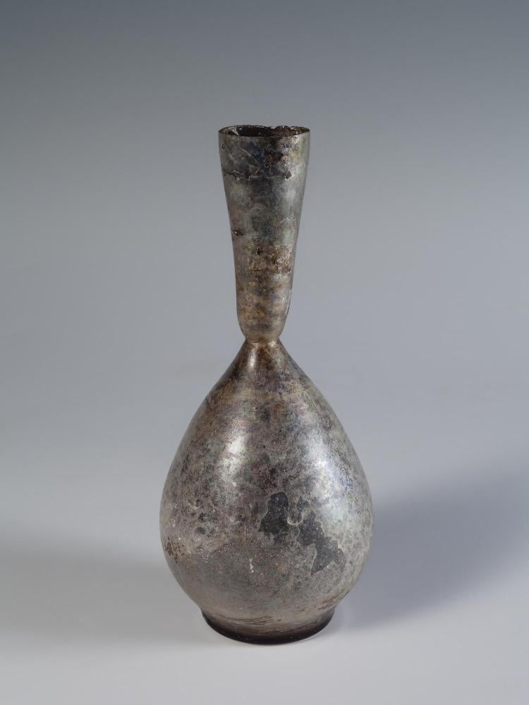 Photograph of a glass vessel with a teardrop-shaped body and a narrow neck with straight, outward tapering sides, from the side against a neutral gray background.