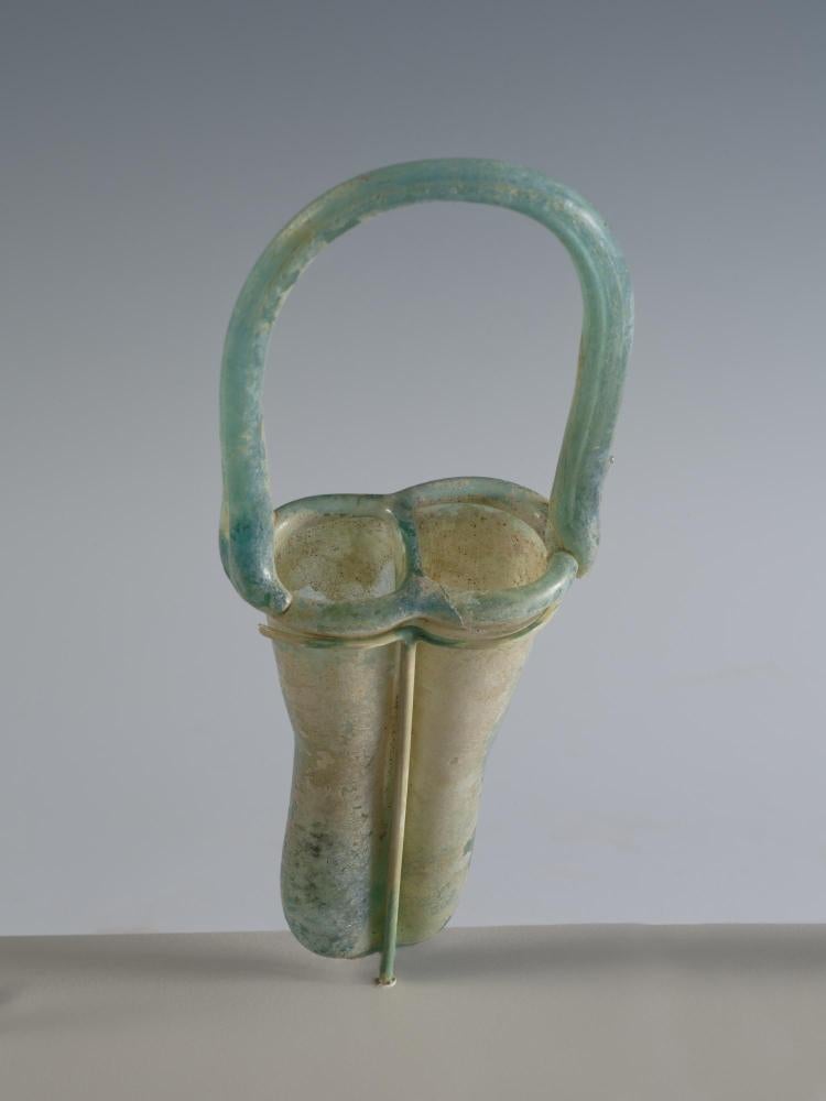 Photograph of a glass unguentarium with two tall cylinders with rounded bottoms and a handle that arches above and across the cylinders, angled toward viewer so interior of cylinders are partially visible, against neutral gray background.