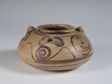 Photograph of a three-handled squat jar, buff clay with brown decoration, from the side against a neutral gray background.