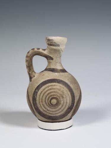 Photograph of a small, rounded jug, tan with brown decoration, against a neutral gray background.