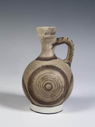 Photograph of a Cypriot juglet with light buff clay and brown decoration, from the side against a neutral gray background.