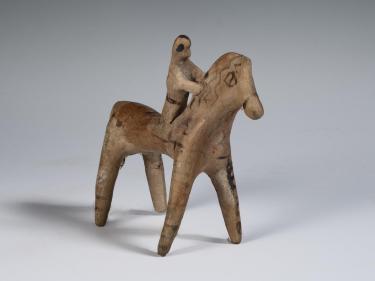 Photograph of horse and rider figurine, from a frontal angle, as if towards viewer's right shoulder, against neutral gray background.