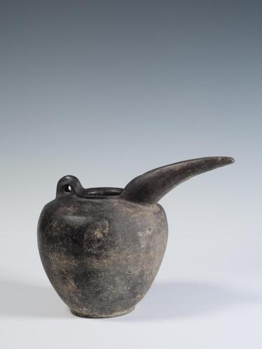 Photograph of grey-ware spouted jug, glossy black covers its entirety, from the side against neutral gray background.