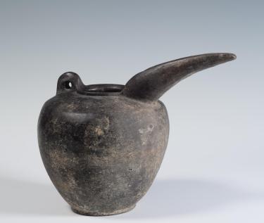 Photograph of a greyware beak-spouted jug, from the side against a neutral gray background.