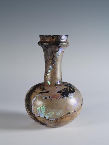 Photograph of a dark glass vessel with a globular body and somewhat tall neck that tapers out at the top, from the side against a neutral gray background.