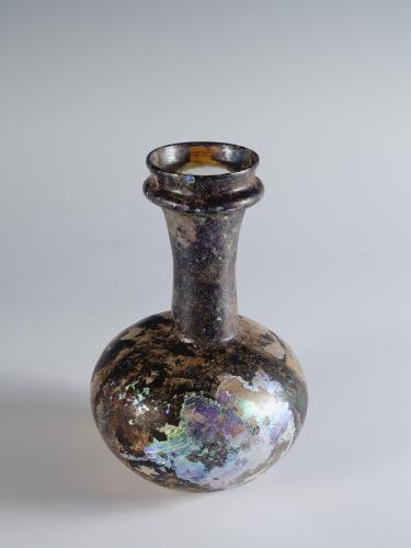 Photograph of a dark glass vessel with a globular body and somewhat tall neck that tapers out at the top, from a slightly raised angle against a neutral gray background.