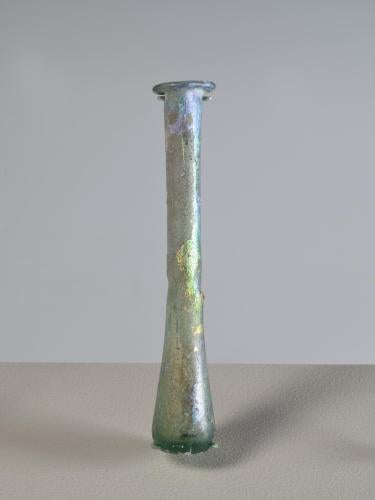 Photograph of a clear glass vessel with a flat bottom, a narrow body and a tall, narrow neck, with a small, rounded mouth, from the side against a neutral gray background.