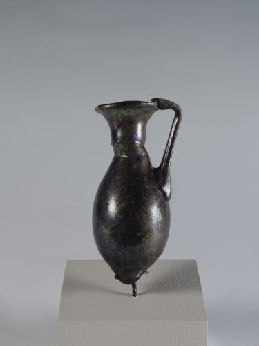 Photograph of a dark glass vessel with an oviod body that tapers to a point, a relatively short neck that tapers outward to the mouth, and a handle that connects the body to the mouth, from the side against a neutral gray background. 