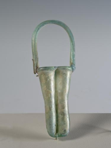 Photograph of a glass unguentarium with two tall cylinders with rounded bottoms and a handle that arches above and across the cylinders, from the side against a neutral gray background.