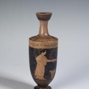 Photograph of red-figure lekythos against a neutral gray background, from the side and showing figure of a woman holding a dish over a wool basket.