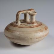 Photograph of stirrup jar, tan with reddish-brown slip decoration, from side angle against neutral gray background.