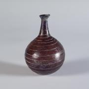 Photograph of a reddish glass vessel with a round body and base with a relatively short and narrow neck and outward-tapering mouth, from the side against a neutral gray background.
