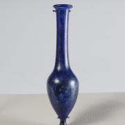 Photograph of a blue glass vessel with a pointed bottom, ovoid body, and tall, narrow neck that tapers slightly outward to a rounded mouth, from the side against a neutral background.