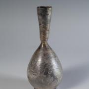 Photograph of a glass vessel with a teardrop-shaped body and a narrow neck with straight, outward tapering sides, from the side against a neutral gray background.