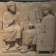 carving of a greek tutor teaching a pupil, Carole Raddato from FRANKFURT, Germany, CC BY-SA 2.0 <https://creativecommons.org/licenses/by-sa/2.0>, via Wikimedia Commons