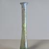 Photograph of a clear glass vessel with a flat bottom, a narrow body and a tall, narrow neck, with a small, rounded mouth, from the side against a neutral gray background.