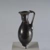 Photograph of a dark glass vessel with an oviod body that tapers to a point, a relatively short neck that tapers outward to the mouth, and a handle that connects the body to the mouth, from the side against a neutral gray background.