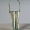 Photograph of a glass unguentarium with two tall cylinders with rounded bottoms and a handle that arches above and across the cylinders, from the side against a neutral gray background.