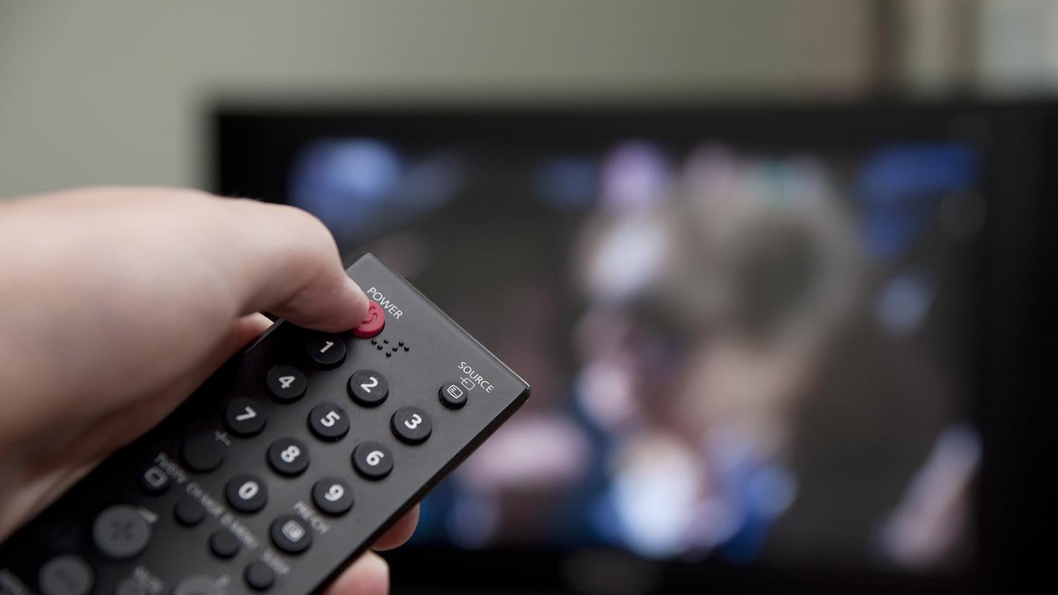 A hand holds a remote control up in front of a TV tuned to the news, intending to hit the power button to turn it off.