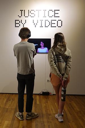 Two young people watch a video. The text Justice By Video is visible in the background.
