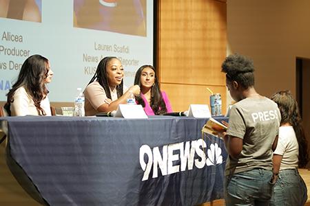 Two students ask journalists questions at a table in a classroom.