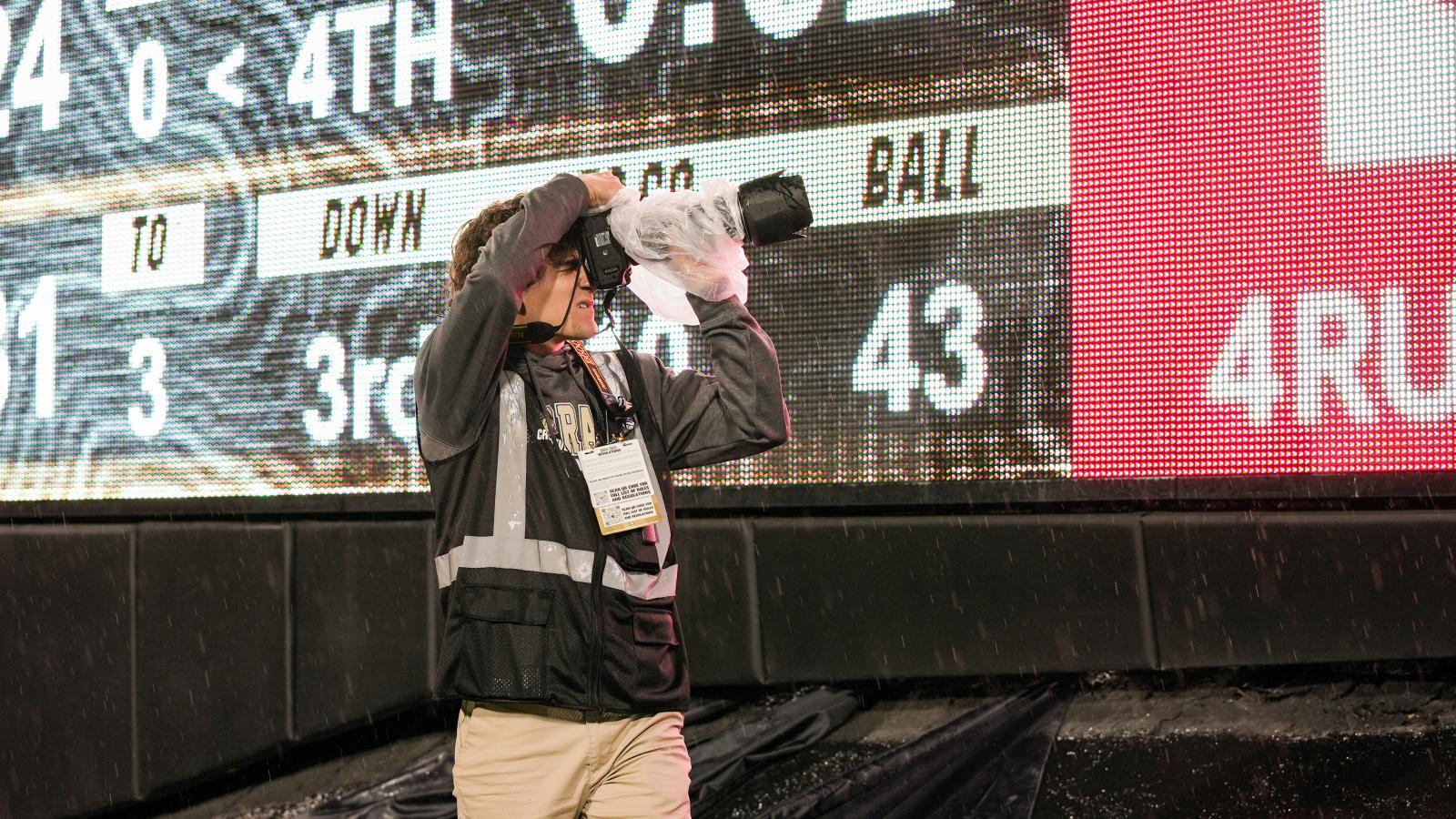 A student uses a camera from the sidelines during a football game. A scoreboard can be seen in the background.