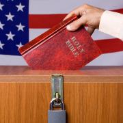 A bible being dropped into a ballot box with an American flag in the background.