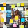A clear board with sticky notes of different colors and writing on it. A business team is visible in the background.