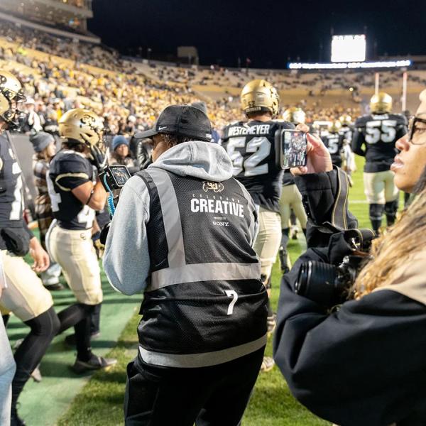 A group of CMCI students shoots video and photos amid a group of players on the sidelines of the stadium during a football game.