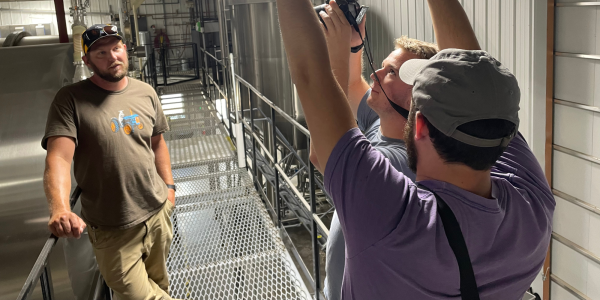 A CPR journalist helps CMCI student William Oster with photo composition while interviewing Todd Olander in his distillery in August. Photo by Paolo Zialcita, CPR
