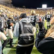 A group of CMCI students shoots video and photos amid a group of players on the sidelines of the stadium during a football game.