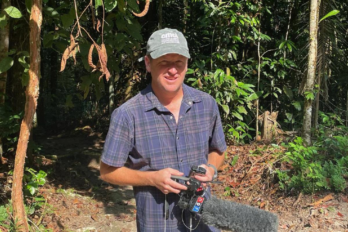 A reporter stands in front of a jungle. He's wearing a CMDI cap and holds and NPR microphone.