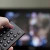 A hand holds a remote control up in front of a TV tuned to the news, intending to hit the power button to turn it off.