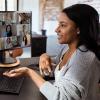 A woman takes a conference call in her home office. Two computer screens are filled with the headshots of participants.