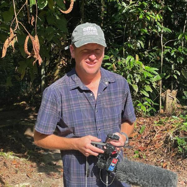 A reporter stands in front of a jungle. He's wearing a CMDI cap and holds and NPR microphone.