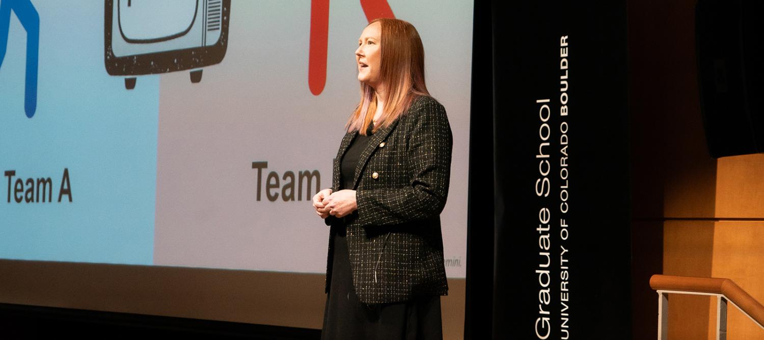 A student in professional attire stands in front of a lecture hall as she defends her dissertation. A slide of her work can be seen in the background.
