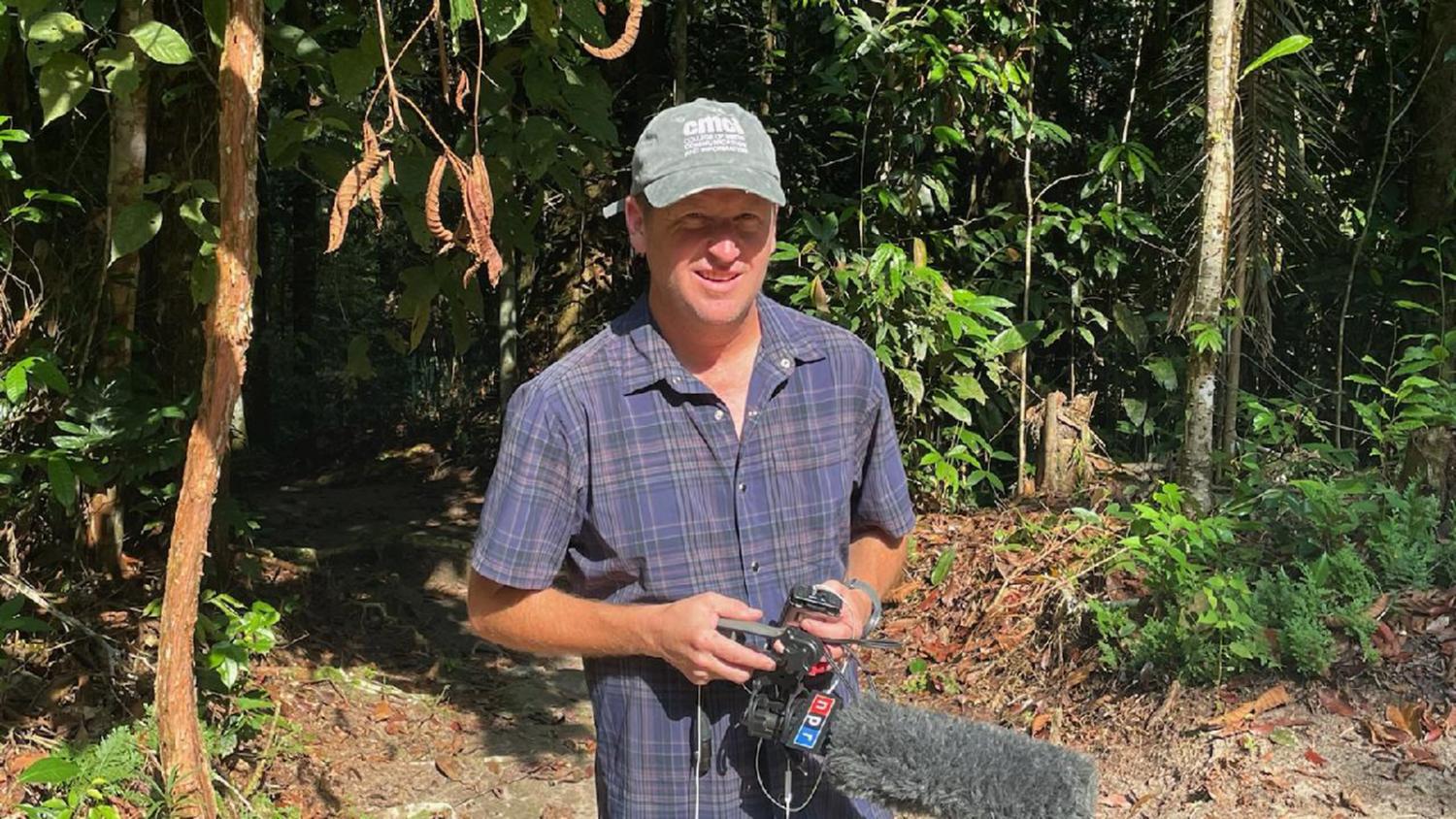A reporter stands in front of a jungle. He's wearing a CMDI cap and holds and NPR microphone.