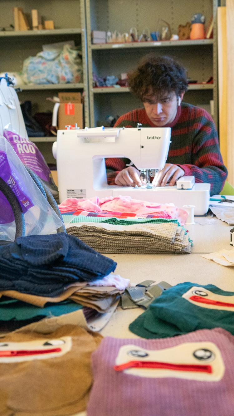 A male student works at a sewing machine, surrounded by different stitched products.